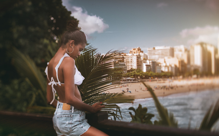 True tilt-shift shot of a young cute black girl checking just filmed photos on her smartphone while standing on the pier with the view on the city beach and the coastline, copy space area on the rightの写真素材