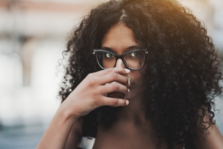 Close-up outdoor portrait of a charming young woman with long curly hair and in glasses drinking delicious coffee from a small glass cup, shallow depth of fieldの写真素材