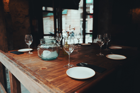 Table setting decor with cutlery, saucers, empty glasses, glass jar with a partially burnt candle inside, wooden table surface; dark interior of a street cafe or cozy restaurant, window behindの写真素材