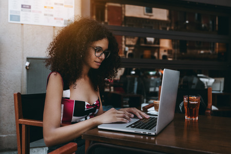 Young beautiful freelancer girl in glasses and with a bulky curly hair is sitting in a street bar and working remotely on her laptop with the glass of delicious cocktail near her on a wooden tableの写真素材