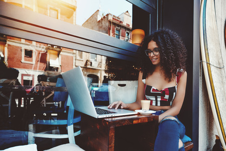 A cheerful young curly caucasian female in eyeglasses is working on her laptop in an outdoor cafe;...