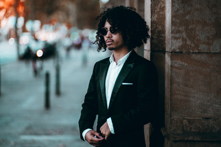 A young elegant Asian man in a formal suit is standing outdoors near a stone pillar; curly stylish Japanese businessman in sunglasses near the column on the street, strong bokeh in the backgroundの写真素材