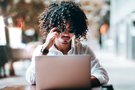 A serious curly Asian man entrepreneur is straightening his round sunglasses while sitting in an outdoor cafe with his laptop and working remotely on his freelance projects; shallow depth of fieldの写真素材