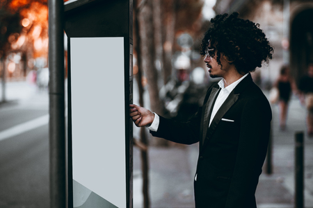 An Asian businessman with curly hair and in sunglasses is paying his parking time via an outdoor terminal; a young man entrepreneur in a formal suit is making payment using parking meter on the streetの写真素材