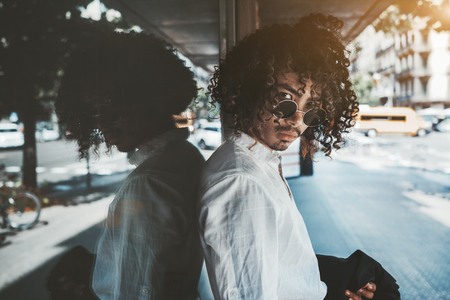 Portrait of a young curly Asian guy in a white shirt and in sunglasses, he is leaning against glass reflecting wall outdoors, looking seriously at the camera, and holding the blazer in his handの写真素材