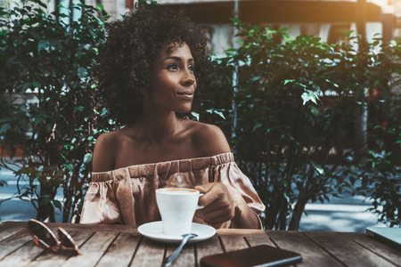 A cheerful young curly female is drinking coffee in a street bar and looking aside; a dazzling happy biracial woman with a cup of tasty hot chocolate is relaxing in a street cafeの写真素材