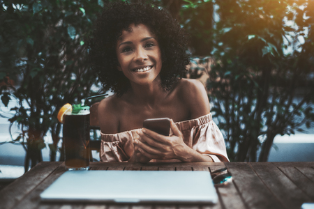A laughing young curly-haired Brazilian female with the smartphone, notebook and a cocktail in a street bar; dazzling happy African-American girl with the cellphone in a street cafe, with a mint drinkの写真素材