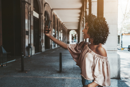 A side view of a curly-hair young African-American female standing in the shadow of the passage between columns of an antique building and taking a selfie using a frontal camera of her smartphoneの写真素材