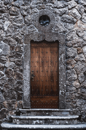 A vertical shot of a beautiful antique wooden riveted door in a stone wall with a small round window...