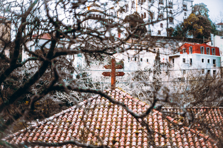 An antique weathered tiled roof with a stone Lorraine cross on it framed with multiple branches of the tree in a defocused foreground, a huge historical building on the hill in the background, Sintraの写真素材