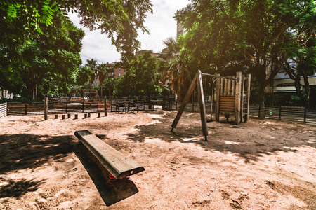 A wide-angle view of an empty contemporary playground surrounded by trees with sand on the ground, wooden bench in the foreground, training apparatus farther, sunny summer day, Barcelona, Spainの写真素材