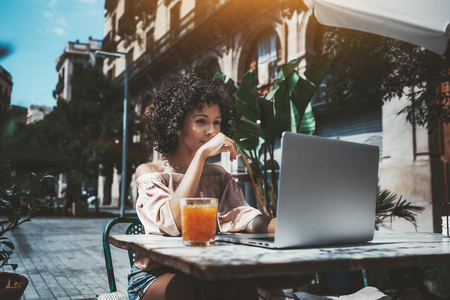 A curly-hair dazzling young African-American female freelancer is working with her project on the laptop remotely from a city cafe outdoors, with a glass of delicious fresh juice near on the tableの写真素材