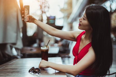 A charming young Asian female with long hair is taking a selfie using her smartphone while sitting in an outdoor bar with a glass of delicious drink and waiting for her friendの写真素材