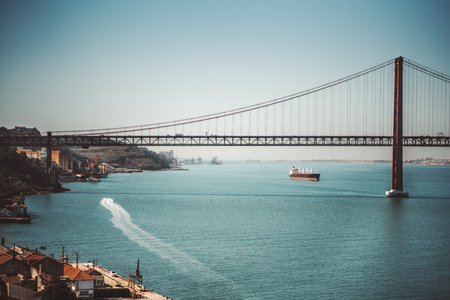 Beautiful scenery with a huge suspension bridge "Ponte 25 de Abril" over the river Tagus in Lisbon, Portugal on a warm sunny day with two vessels, one of them is leaving a long trace on the waterの写真素材
