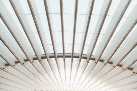 A wide-angle view of a white outdoor roof construction of a modern building made of parallel thick hollow iron girders and translucent covering to pass and scatter the sunlight