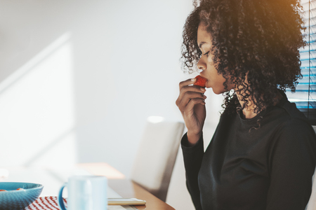 Cute curly-hair African-American girl is eating strawberry during her morning breakfast while sitting at the kitchen and reading the news feed on her smartphone on the table with a cup of coffee nearの写真素材