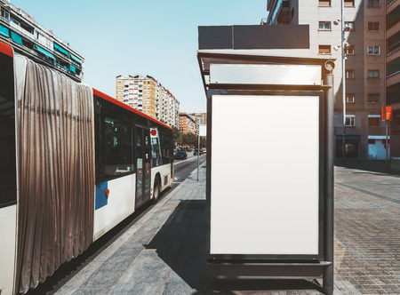 An empty ad poster mock-up on an outer side of the bus stop; advertising billboard placeholder template on a stop of public transport with a bus on the left; mockup of a blank white outdoor bannerの写真素材