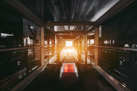 Long dark glass contemporary corridor in an airport terminal departure area or an arrival zone of modern train station depot with a descending moving walkway, and silhouettes of people in the distanceの写真素材