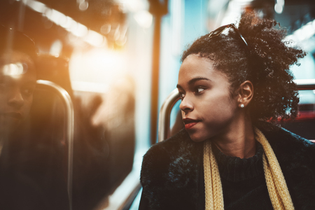 The portrait of a young charming African-American female pensively looking outside the carriage window while sitting indoors of a metro train; Brazilian girl in a subway train, shallow depth of fieldの写真素材