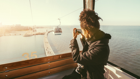 Rear view of a young tourist female shooting evening scenery using a camera of her smartphone while sitting inside of a modern ropeway car with a copy space place on the left for your logo or ad textの写真素材