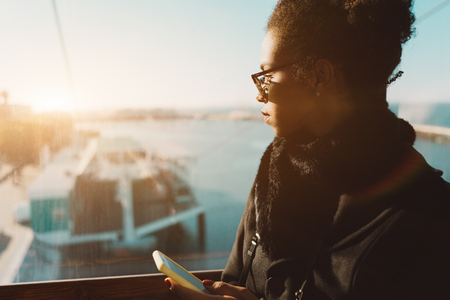 Side view of a young African-American woman with a cellphone inside of ropeway cabin; charming Brazilian girl in sunglasses is typing a message via smartphone while standing next to the windowの写真素材