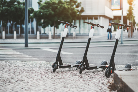 Electric urban transportation: three modern electric readies to ride scooter bikes with accumulators in the center of a city on the pavement stone with the road and crosswalk behind, Lisbon, Portugalの写真素材