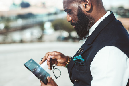 Side view of African bearded stylish man entrepreneur holding a futuristic tablet computer in his hand and implementing a secure login using privacy interface; adult black businessman with the gadgetの写真素材