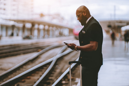 A fancy bald adult African senior with a beard and in glasses is using a map application in his smartphone while waiting for the train on the railway station depot, with a copy space place on the leftの写真素材