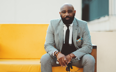 A serious adult bearded bald African man in an elegant plaid costume with a tie is sitting on a yellow soft sofa outdoors and looking at the camera with round spectacles in his handの写真素材