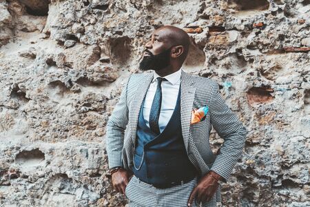 Handsome bald bearded black senior in an elegant costume in front of a grungy wall outdoors; a fashion adult African man with the beard and in a suit with vest and necktie standing near a stone wallの写真素材