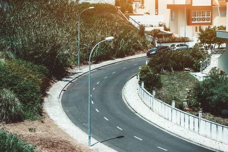 View from the top of a road turn in a residential district of Sintra, Portugal, with road marking in a center, two street lights, dwelling houses with a stone fence on the right, bushes on the leftの写真素材