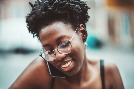 A close-up outdoor portrait of a happy African girl in spectacles during a pleasant conversation with her mom via the smartphone which she holds without hands just holding between cheek and shoulderの写真素材