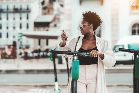Young cheerful elegant African female in eyeglasses and cloak, with a nails art, is using  her smartphone outdoors to activate via internet a rental street e-scooter with a green battery aboveの写真素材