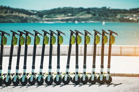 Urban electric transportation outdoors: an unfinished row of slightly shabby electric scooter bikes with green batteries blocks on the top, embankment of Lisbon, Portugal, with the Tagus river behindの写真素材