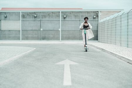 A young African fancy female in glasses and a white fluttering cloak is riding an e-scooter on the asphalt road with a painted arrow as a road marking in a defocused foreground, shallow depth of fieldの写真素材