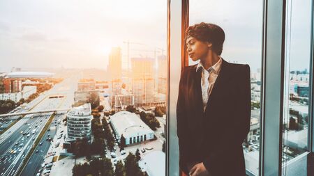 A pensive African-American woman entrepreneur is thoughtfully looking at the cityscape outside the...