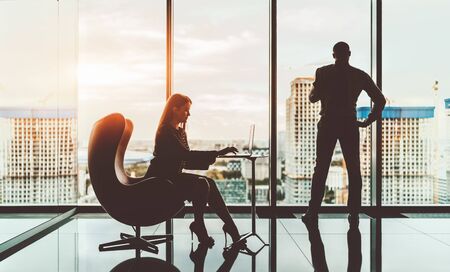 Silhouettes of two business colleagues: a businesswoman is sitting on an armchair and using a laptop and a man entrepreneur standing next to the window and looking at cityscape and construction siteの写真素材