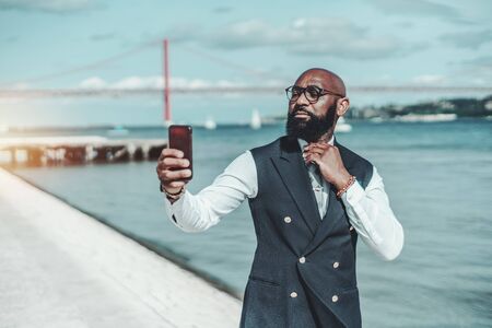 An elegant African man in glasses is taking a selfie on cellphone while standing near the river; a stylish black guy in spectacles is using a camera of his smartphone as a mirror to straighten his tieの写真素材