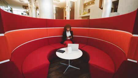 Wide-angle view of an African-American woman entrepreneur sitting inside of the area of a red round sofa in an office with a small white table in the center and a laptop on it and using her smartphoneの写真素材