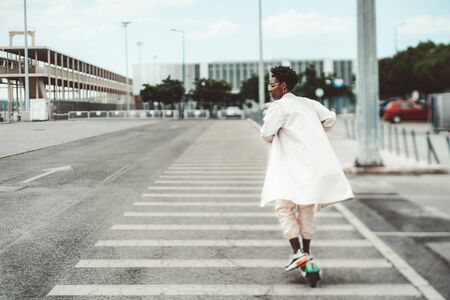 True tilt-shift shot of a young African female in glasses and a white fluttering cloak riding an e-scooter on a wide road with marking on it on a cloudy dayの写真素材