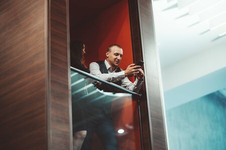 A man entrepreneur is showing a message on the screen of his smartphone to his female colleague while they both standing on the balcony in a modern luxury office lounge area and having a business chatの写真素材