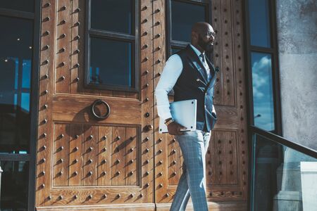 An elegant bald bearded African man entrepreneur in eyeglasses and vest has just left a modern building after a business meeting and coming downstairs while carrying a laptop in his handの写真素材