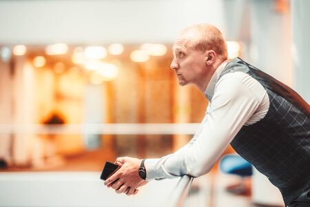 Sideview of a confident handsome adult man entrepreneur leaning against a chromium railing in the office open space area and pensively looking into the distance while holding a smartphone in his handsの写真素材