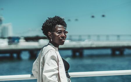 Portrait of a young charming African female in sunglasses and white trench standing on the quayside with a ropeway in the defocused background; black girl on the pier with cableway behind herの写真素材