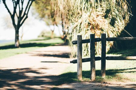 Fragment of a wooden fence made of thick round girders in the foreground on a warm sunny day in a park with a pathway in the center of the frame and a tree in the background balancing the compositionの写真素材