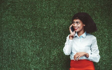 A smiling young African-American businesswoman  is phoning in front of a green grassy wall in an office; a charming cheerful biracial woman entrepreneur is speaking on the smartphone indoorsの写真素材