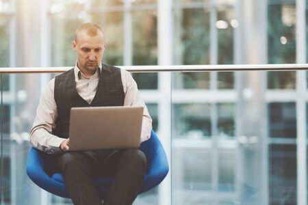 A serious pensive mature man entrepreneur is sitting on a blue armchair in office open-space settings and using his laptop, with a copy space area on the right for a logo or an advertising messageの写真素材