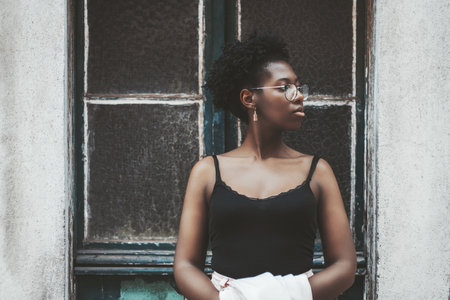 A charming young African female in spectacles is standing in front of an old antique wall with a doorway and  pensively looking aside; a charming black girl in eyeglasses outdoors near the doorの写真素材