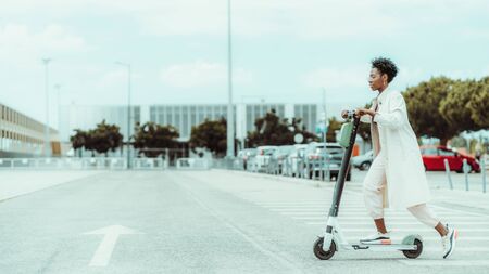 Young fancy African woman in spectacles and a white cloak is riding an e-scooter with green battery above on the asphalt road with a painted arrow as a road marking, sunny day, shallow depth of fieldの写真素材