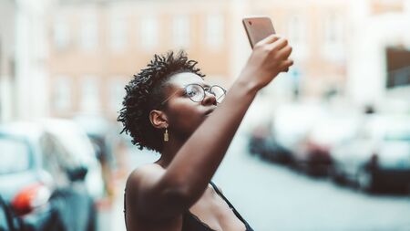 Portrait of a cute African girl in spectacles taking a photo of a showplace using a smartphone; beautiful young black female in eyeglasses is photographing something above the street on her cellphoneの写真素材